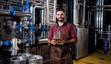 Modern bira fabrikasında bira fermantasyonunu denetleyen deri önlüklü genç bir erkek bira üreticisi.