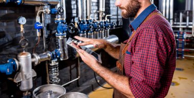 Modern bira fabrikasında bira fermantasyonunu denetleyen deri önlüklü genç bir erkek bira üreticisi.