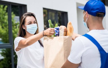 Koruyucu maske takan ve eldiven takan genç bir kurye karantina sırasında genç bir kadına mal teslim ediyor.