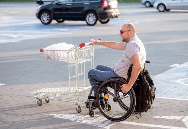 Tekerlekli sandalyedeki engelli adam süpermarketin önünde arabasını itiyor.