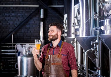 Deri önlük giyen genç bira imalathanesinde bira test ediyor.