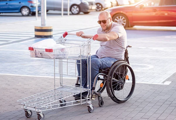 Tekerlekli sandalyedeki engelli adam süpermarketin önünde arabasını itiyor. — Stok fotoğraf