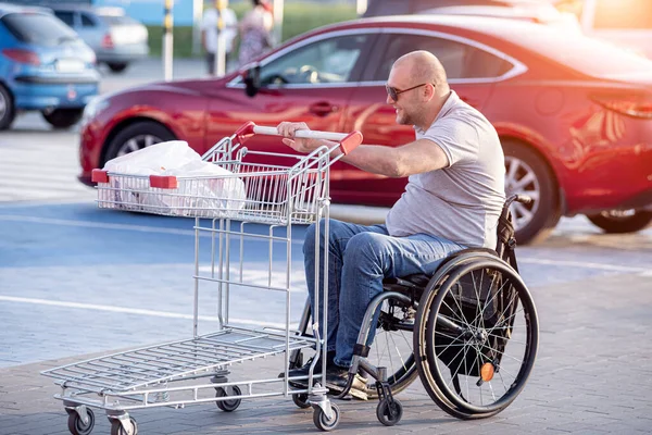 Tekerlekli sandalyedeki engelli adam süpermarketin önünde arabasını itiyor. — Stok fotoğraf
