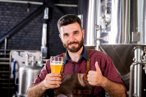 Jovem cervejeiro vestindo um avental de couro está testando cerveja em uma cervejaria moderna — Fotografia de Stock