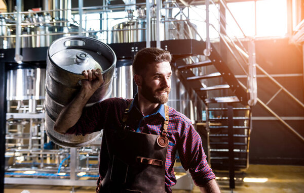 Young male brewer in leather apron holds barrel with craft beer at modern brewery factory