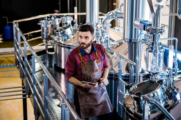 Jovem cervejeiro macho em avental de couro supervisionando o processo de fermentação da cerveja na fábrica moderna de cervejaria — Fotografia de Stock