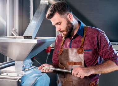 Deri önlüklü genç bir bira üreticisi modern bir bira fabrikasında malt tohumlarının öğütülmesini kontrol eder.