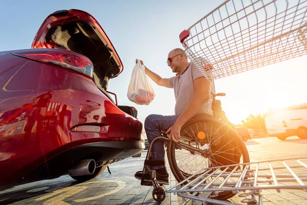 Vuxna funktionshindrade man i rullstol lägger inköp i bagageluckan på en stormarknad parkeringsplats — Stockfoto