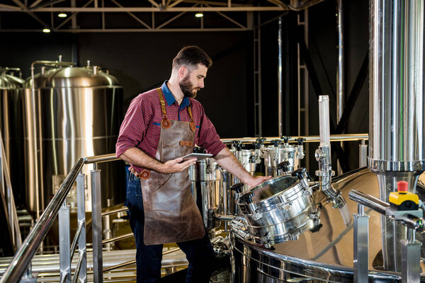 Молодой пивовар в кожаном фартуке контролирует процесс брожения пива на современном пивоваренном заводе