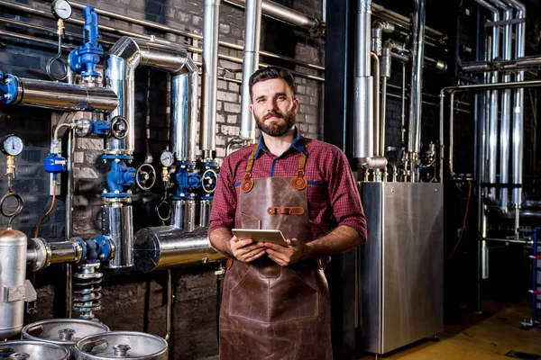 Jovem cervejeiro macho em avental de couro supervisionando o processo de fermentação da cerveja na fábrica moderna de cervejaria — Fotografia de Stock