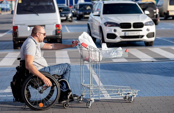 Tekerlekli sandalyedeki engelli adam süpermarketin önünde arabasını itiyor. — Stok fotoğraf