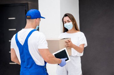 Koruyucu maske takan ve eldiven takan genç bir kurye karantina sırasında genç bir kadına mal teslim ediyor.