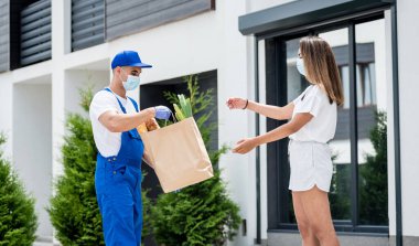 Koruyucu maske takan ve eldiven takan genç bir kurye karantina sırasında genç bir kadına mal teslim ediyor.