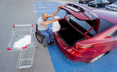 Tekerlekli sandalyedeki yetişkin bir adam süpermarketin otoparkındaki bir arabanın bagajına alışveriş yapıyor.