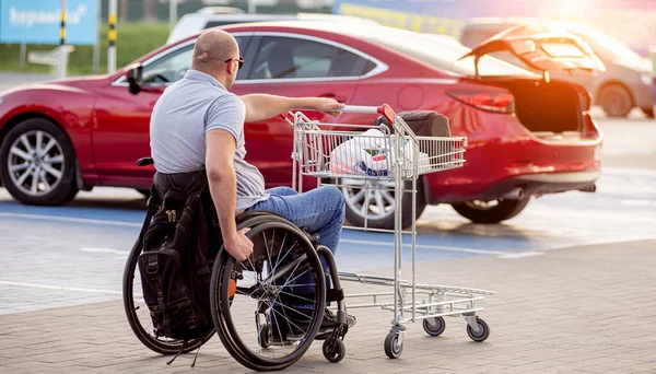 Vuxen handikappad man i rullstol kör en vagn mot en bil på en stormarknad parkeringsplats — Stockfoto