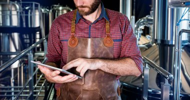 Modern bira fabrikasında bira fermantasyonunu denetleyen deri önlüklü genç bir erkek bira üreticisi.