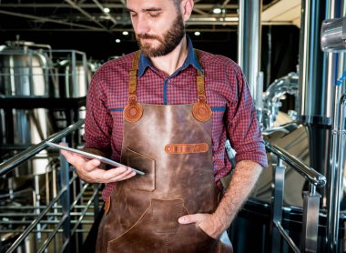 Modern bira fabrikasında bira fermantasyonunu denetleyen deri önlüklü genç bir erkek bira üreticisi.