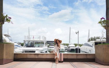 Pembe bavullu bir kadın, yat limanının önündeki beton bir çitin üzerinde oturuyor..