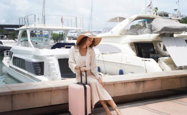 Pembe bavullu bir kadın, yat limanının önündeki beton bir çitin üzerinde oturuyor..