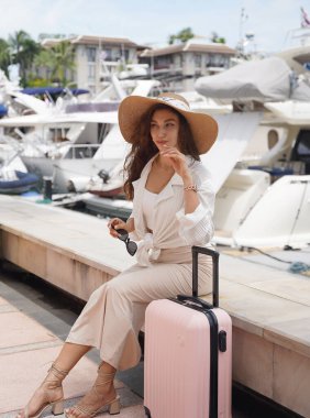 Pembe bavullu bir kadın, yat limanının önündeki beton bir çitin üzerinde oturuyor..