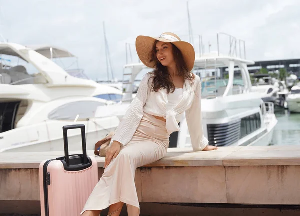 Pembe bavullu bir kadın, yat limanının önündeki beton bir çitin üzerinde oturuyor..