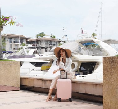 Pembe bavullu bir kadın, yat limanının önündeki beton bir çitin üzerinde oturuyor..