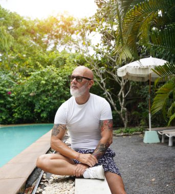 Bir adam tropikal bölgede bir yüzme havuzunun yanında rahatça oturuyor..