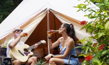 Bir erkek ve bir kadın neşeyle bira içiyor ve glamping 'de rahat bir çadırın yanında gitar çalıyorlar..