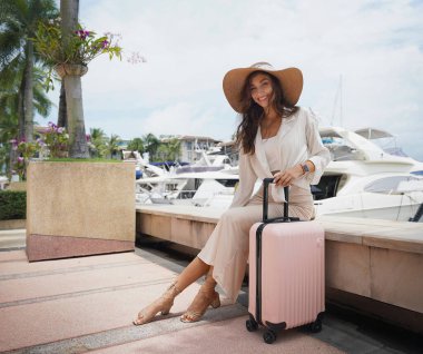 Pembe bavullu bir kadın, yat limanının önündeki beton bir çitin üzerinde oturuyor..