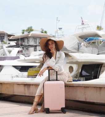 Pembe bavullu bir kadın, yat limanının önündeki beton bir çitin üzerinde oturuyor..