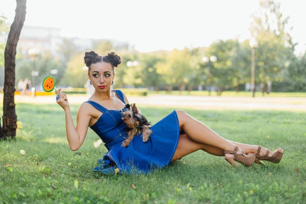 Bright makeup beautiful girl with Yorkshire Terrier seating on the grass holding watermelon lollipop.