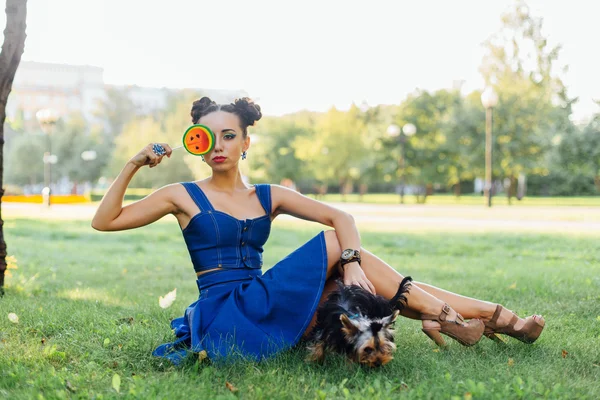 Ljusa makeup vacker flicka med Yorkshire Terrier sittplatser på gräset håller watermelon lollipop. — Stockfoto