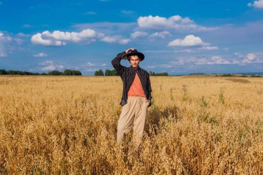 Kırsal Kır Sahnesi. Siyah gömlekli ve siyah şapkalı uzun boylu yakışıklı adam altın yulaf tarlasında duruyordu. Mavi gökyüzü ile yaz manzarası