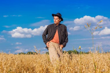 Kırsal Kır Sahnesi. Siyah gömlekli ve siyah şapkalı uzun boylu yakışıklı adam altın yulaf tarlasında duruyordu. Mavi gökyüzü ile yaz manzarası