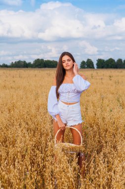 Kırsal Kır Sahnesi. Altın yulaf tarlasında beyaz giysili, uzun saçlı genç bir kadın yulaf kulaklı sepeti tutuyor..