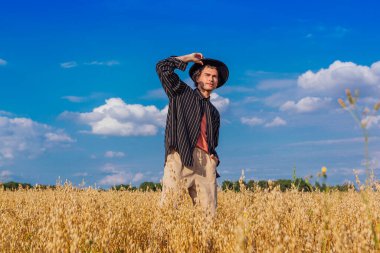 Kırsal Kır Sahnesi. Siyah gömlekli ve siyah şapkalı uzun boylu yakışıklı adam altın yulaf tarlasında duruyordu. Mavi gökyüzü ile yaz manzarası