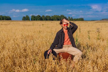Kırsal Kır Sahnesi. Siyah gömlekli, siyah şapkalı ve pembe güneş gözlüklü uzun boylu yakışıklı adam altın yulaf tarlasında kahverengi bir deri valizde oturuyor. Mavi gökyüzü ile yaz manzarası