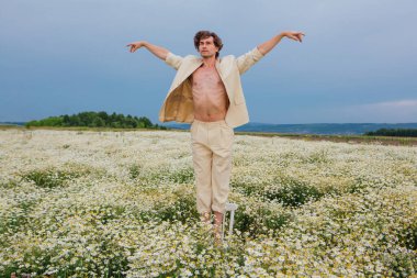 Uzun boylu, beyaz takım elbiseli, çıplak vücutlu, papatya çiçeği tarlasında beyaz sandalyede dans eden yakışıklı bir adam.