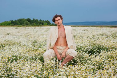 Beyaz takım elbiseli uzun boylu, yakışıklı bir adam papatya çiçekleri tarlasında bir sandalyenin arkasında oturuyordu.