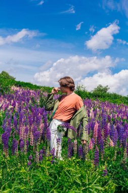 Yeşil ceketli uzun boylu yakışıklı bir adam lupin çiçeği tarlasında duruyor cebinde lupin çiçekleri, doğanın güzelliğini emrediyor. Mor ve pembe lupinlerle çevrili bir adam..