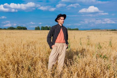 Kırsal Kır Sahnesi. Siyah gömlekli ve siyah şapkalı uzun boylu yakışıklı adam altın yulaf tarlasında duruyordu. Mavi gökyüzü ile yaz manzarası