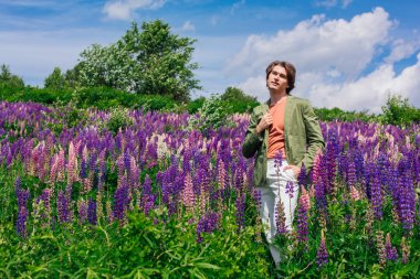Yeşil ceketli uzun boylu yakışıklı bir adam lupin çiçeği tarlasında duruyor, doğanın güzelliğini emrediyor. Mor ve pembe lupinlerle çevrili bir adam..