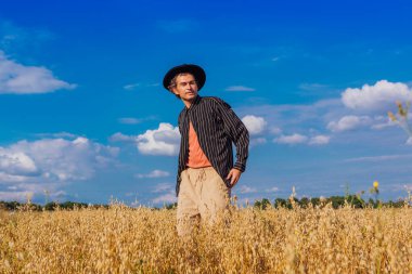 Kırsal Kır Sahnesi. Siyah gömlekli ve siyah şapkalı uzun boylu yakışıklı adam altın yulaf tarlasında duruyordu. Mavi gökyüzü ile yaz manzarası