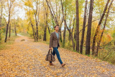 Kahverengi ceketli uzun boylu yakışıklı bir adam sonbahar sokağında çantasıyla yürüyor.