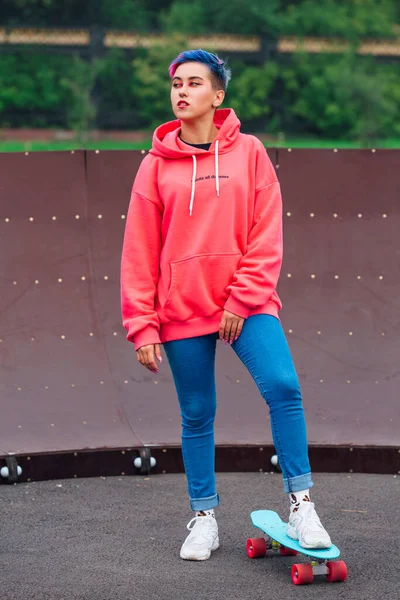 Portrait of a trendy pretty young girl dressed in pink hoodie and jeans standing next to the skateboard court with her blue plastic skateboard.