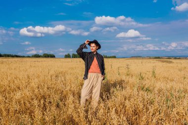 Kırsal Kır Sahnesi. Siyah gömlekli ve siyah şapkalı uzun boylu yakışıklı adam altın yulaf tarlasında duruyordu. Mavi gökyüzü ile yaz manzarası