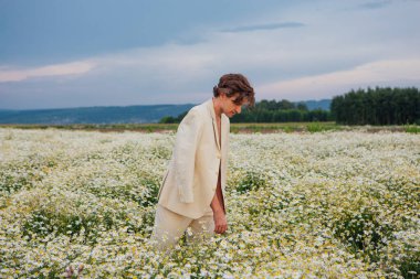 Uzun boylu, beyaz takım elbiseli, çıplak vücutlu yakışıklı bir adam papatya çiçeği tarlasında dikiliyor.