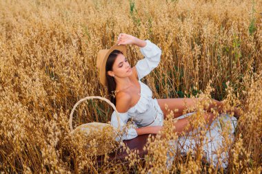 Kırsal Kır Sahnesi. Beyaz bluz ve hasır şapka giymiş, uzun saçlı genç ve güzel bir kadın altın yulaf tarlasında, yulaf kulaklı sepeti tutuyordu..