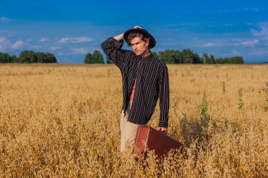 Kırsal Kır Sahnesi. Siyah gömlekli ve siyah şapkalı uzun boylu yakışıklı adam altın yulaf tarlasında kahverengi deri valiziyle dikiliyordu. Mavi gökyüzü ile yaz manzarası