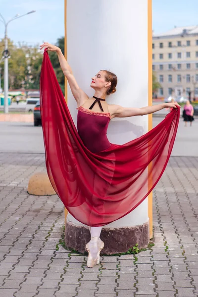 Woman ballerina in red ballet dress dancing in pointe shoes next to the old columns. Ballerina standing in beautiful ballet pose